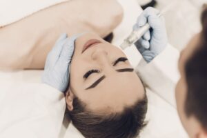 Woman receiving a professional facial treatment with a microneedling, performed by a gloved specialist in a clinical setting.