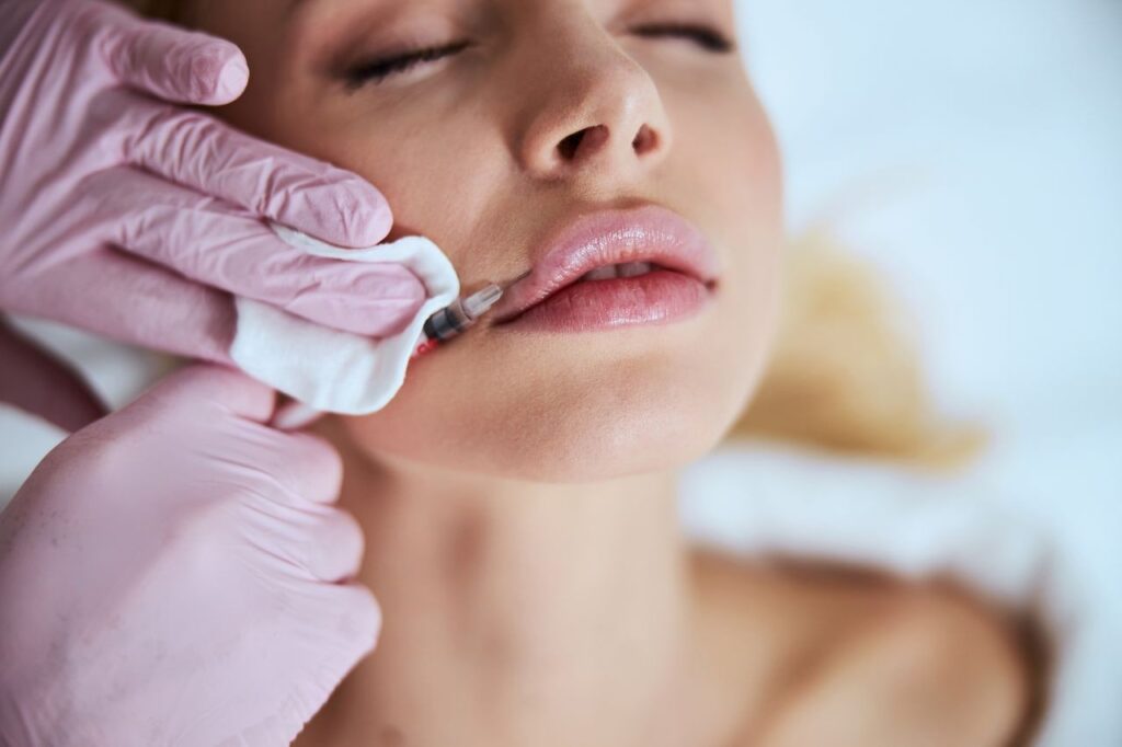 A close-up of a woman receiving a lip injection procedure, with a medical professional wearing pink gloves performing the treatment using a syringe.